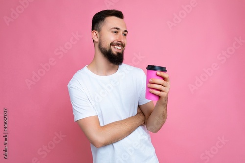 Handsome positive young brunette bearded man wearing white t-shirt isolated over pink backgroung wall holding paper coffee cup drinking and looking to the side