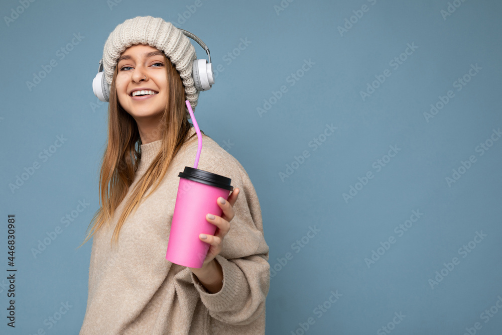 Photo shot of Beautiful joyful smiling young blond woman wearing beige winter sweater and hat isolated over blue background drinking smoothie wearing white wireless bluetooth headphones listening to Photo shot of Beautiful joyful smiling young blond woman wearing beige winter sweater and hat isolated over blue background drinking smoothie wearing white wireless bluetooth headphones listening to