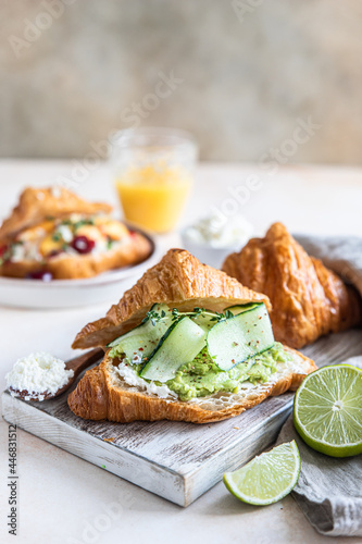 Croissant sandwich with ricotta or cream cheese, mashed avocado and cucumber on wooden cutting board. Delicious brunch or breakfast.