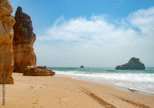 Empty clean beach, slightly orange sand with tire tracks, cliffs in the background, ocean waves and clouds in the sky