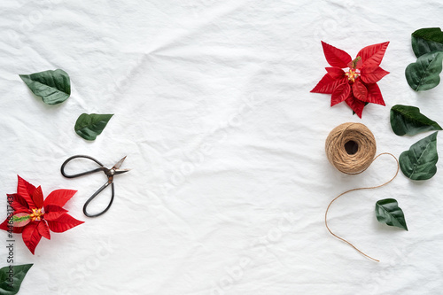 Christmas flat lay with copy-space. Red poinsettia, Xmas top view on off white, ivory textile background, text space. Hemp cord, scissors, red and dark green poinsettia leaves . © tilialucida