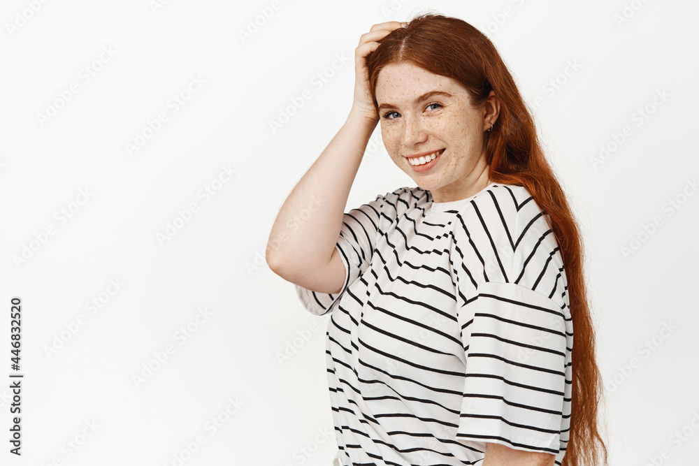 Natural beauty. Girl with red hair and freckles, touch her head, smiling happy and gazing at camera, confident and cheerful face expression, posing in white studio against background Natural beauty. Girl with red hair and freckles, touch her head, smiling happy and gazing at camera, confident and cheerful face expression, posing in white studio against background