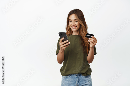 Smiling adult woman paying online, holding credit discount card and looking happy at mobile phone screen, shopping in internet, using app to purchase, white background