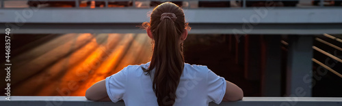 unrecognizable teenage girl with long hair in ponytail admires setting sunset in orange sky standing on overpass bridge backside upper view