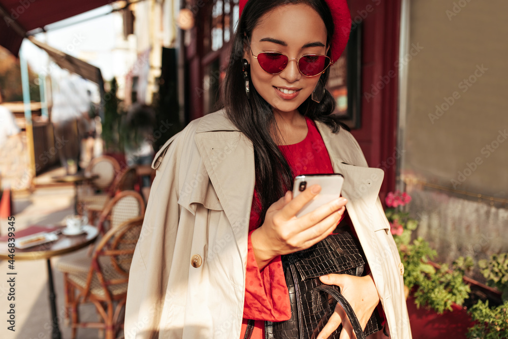 Attractive brunette Asian lady in beige trench coat, red dress, beret and eyeglasses messaging in phone and holds black handbag.