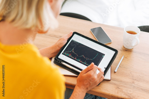 Young blonde woman in yellow t-shirt studies cryptocurrency charts and stock quotes on digital tablet with cup of tea in kitchen at home