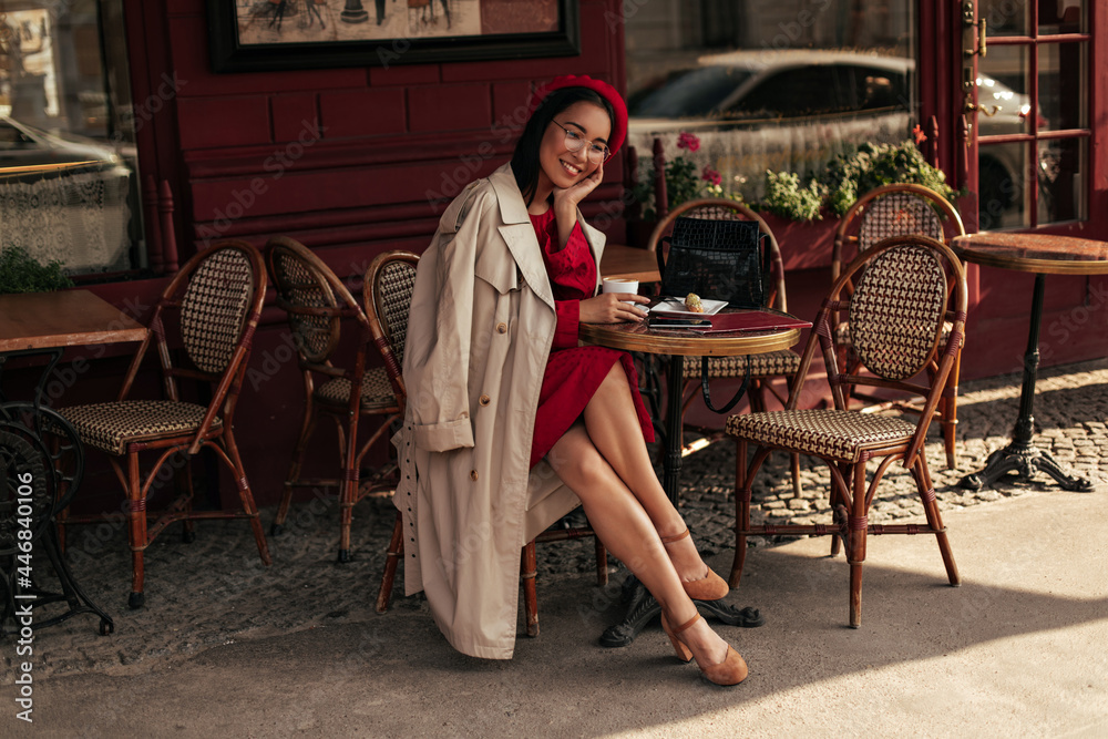 Pretty Asian brunette lady in red dress and beige trench coat smiles sincerely, sits in street cafe and holds coffee cup. Charming woman in beret and eyeglasses poses in restaurant. Pretty Asian brunette lady in red dress and beige trench coat smiles sincerely, sits in street cafe and holds coffee cup. Charming woman in beret and eyeglasses poses in restaurant.