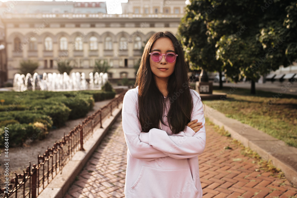 Fototapeta premium Serious brunette Asian woman in pink sunglasses and stylish hoodie crosses arms and looks into camera outside.