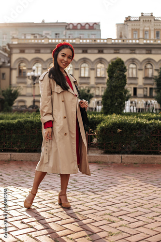 Full-length portrait of brunette woman in beige trench coat, red beret and dress smiles widely and holds handbag outside. © Look!