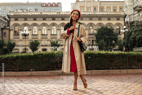 Elegant young brunette woman in stylish beige trench coat, red midi dress and beret smiles and poses in good mood outside.