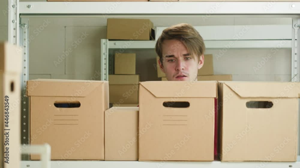 Man wearing red uniform T-shirt, looking for parcel and taking it, employee of warehouse finding delivery box on the background of cardboard drawers. Logistics, delivering and storage concept. 