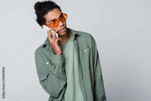african american guy in shirt and eyeglasses talking on smartphone isolated on grey