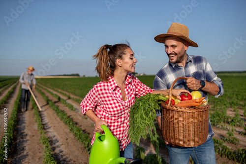 Young couple holding watering can and basket with vegetables and elderly farmer working in background.