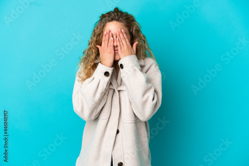 Young blonde woman isolated on blue background covering eyes by hands