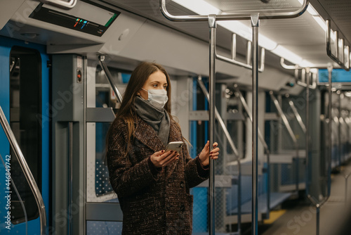 A woman in a face mask is standing and using a smartphone in a modern subway car
