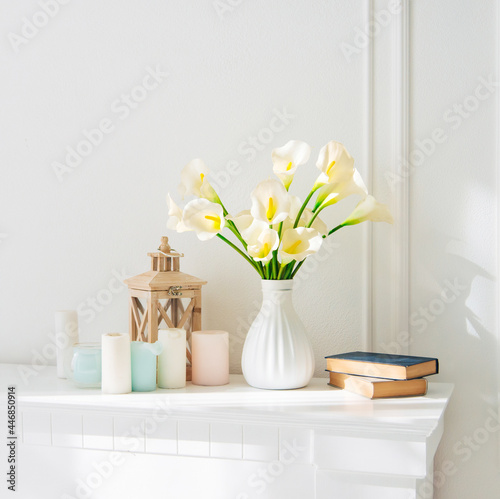 Soft home decor, white jug, vase with white and yellow beautiful flowers on a white wall background and on a wooden shelf. Interior. 