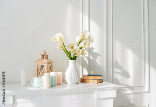 Soft home decor, white jug, vase with white and yellow beautiful flowers on a white wall background and on a wooden shelf. Interior.