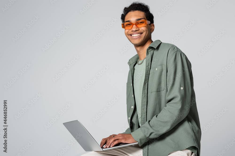 cheerful african american guy in stylish eyeglasses typing on laptop and smiling at camera on grey background