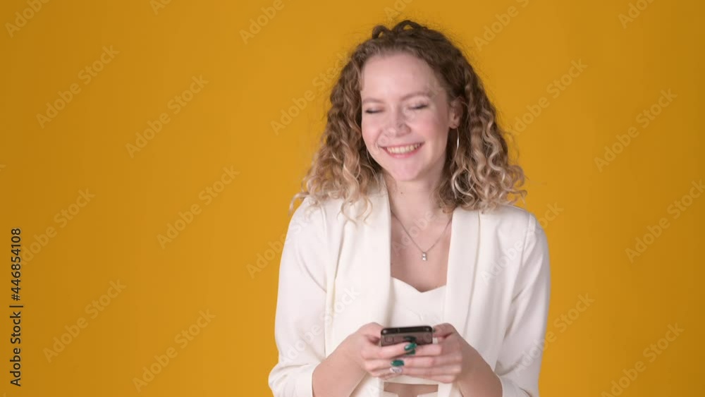 Happy young girl with curly hair using smart phone and texting online chat with smile isolated on yellow background. People lifestyle concept. Happy young girl with curly hair using smart phone and texting online chat with smile isolated on yellow background. People lifestyle concept.
