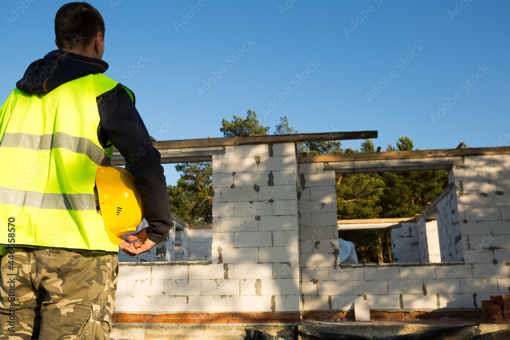 © Ольга Симонова - Builder holds in hand a yellow protective hardhat on the construction site with the foundation and walls of the house made of cellular concrete blocks and construction materials. Work safety, mockup © Ольга Симонова - Builder holds in hand a yellow protective hardhat on the construction site with the foundation and walls of the house made of cellular concrete blocks and construction materials. Work safety, mockup