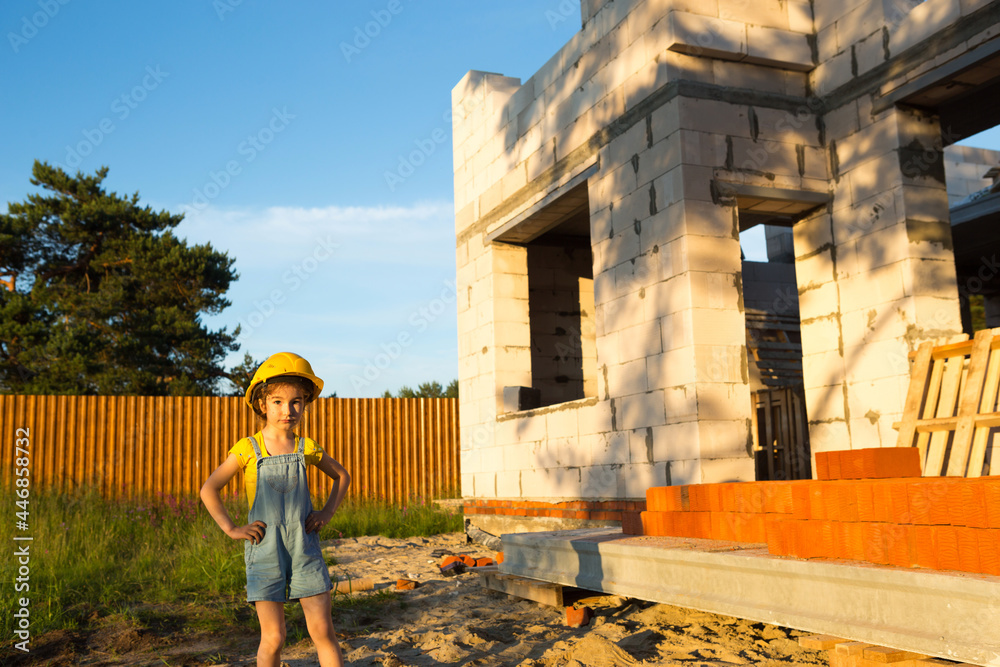 A little girl in a yellow hardhat is playing builder on the construction site of her future home. Dreams of moving, choosing a profession, children's education.