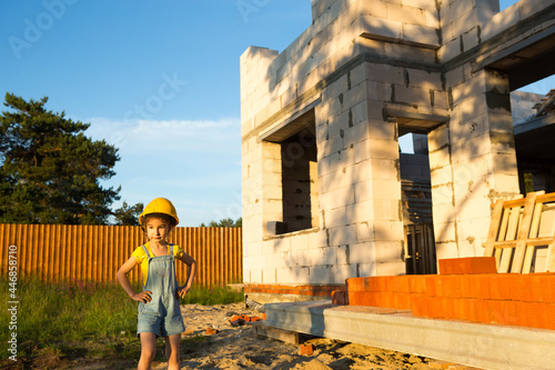 A little girl in a yellow hardhat is playing builder on the construction site of her future home. Dreams of moving, choosing a profession, children's education.
