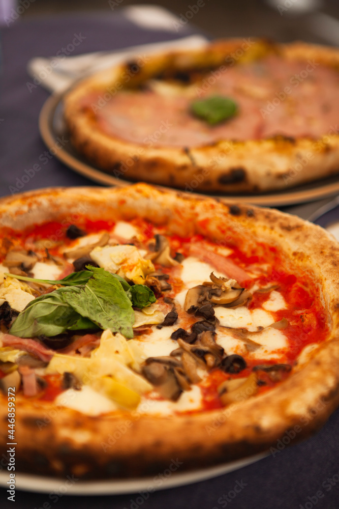 Close up. Two traditional Italian round pizzas with prosciutto, mushrooms, ham, pistachio, olives, basil and mozzarella cheese at the restaurant in Milan, Lombardy, Italy. European food and snacks. Close up. Two traditional Italian round pizzas with prosciutto, mushrooms, ham, pistachio, olives, basil and mozzarella cheese at the restaurant in Milan, Lombardy, Italy. European food and snacks.