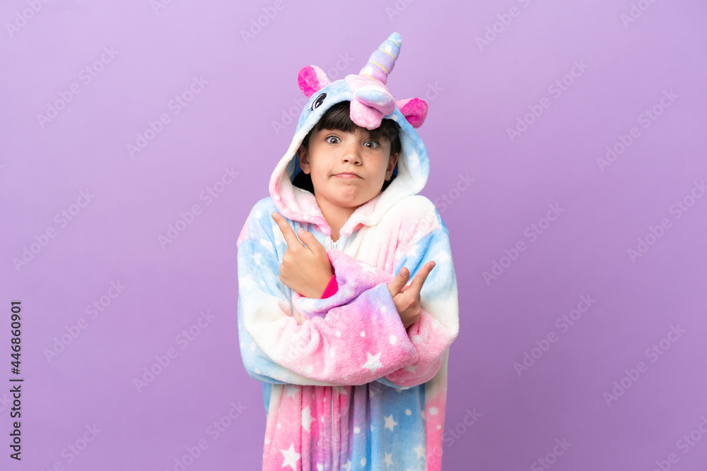 Little kid wearing a unicorn pajama isolated on purple background pointing to the laterals having doubts