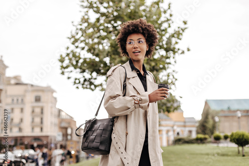Charming young brunette curly woman in beige trench coat holds leathered black handbag and coffee glass outside.