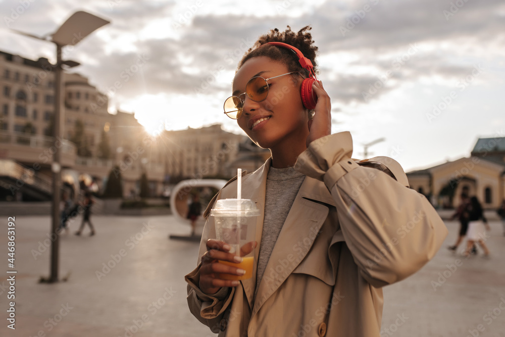 Fototapeta premium Cheerful woman in beige trench coat and eyeglasses enjoys listening to music in headphones outside. Lady drinks juice during sunset.