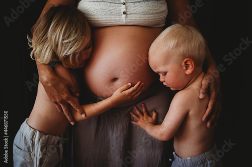 Young blonde siblings hugging mom's pregnant big belly at home photo