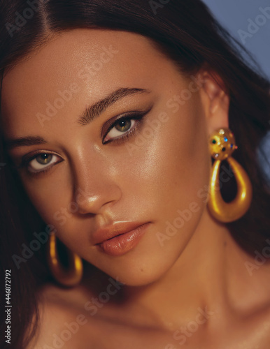 Beautiful woman smooth long hair brunette evening make up with liner tanned skin beautiful female portrait over blue background wearing gold earrings