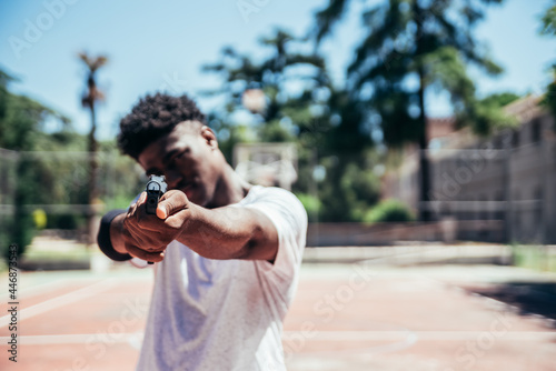 Black African American boy on a basketball court aiming a gun at camera. Focus on the gun. photo
