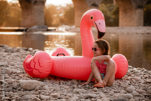 Girl sits in front of flamingo float next to river in front of bridge photo