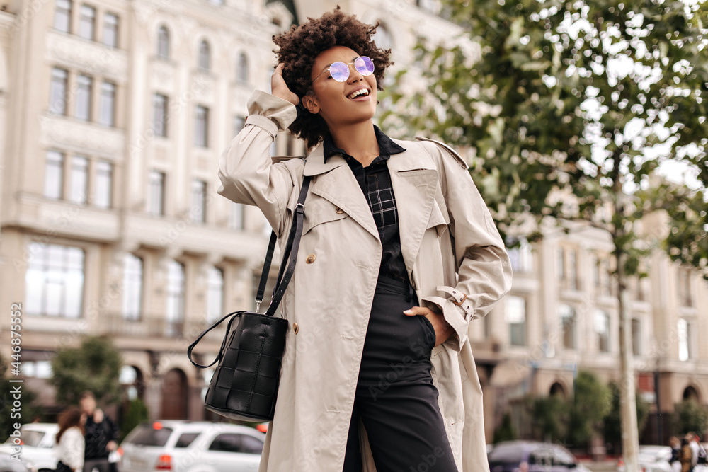 Stylish happy woman in black pants and blouse, beige trench coat and eyeglasses touches hair and walks outdoors.