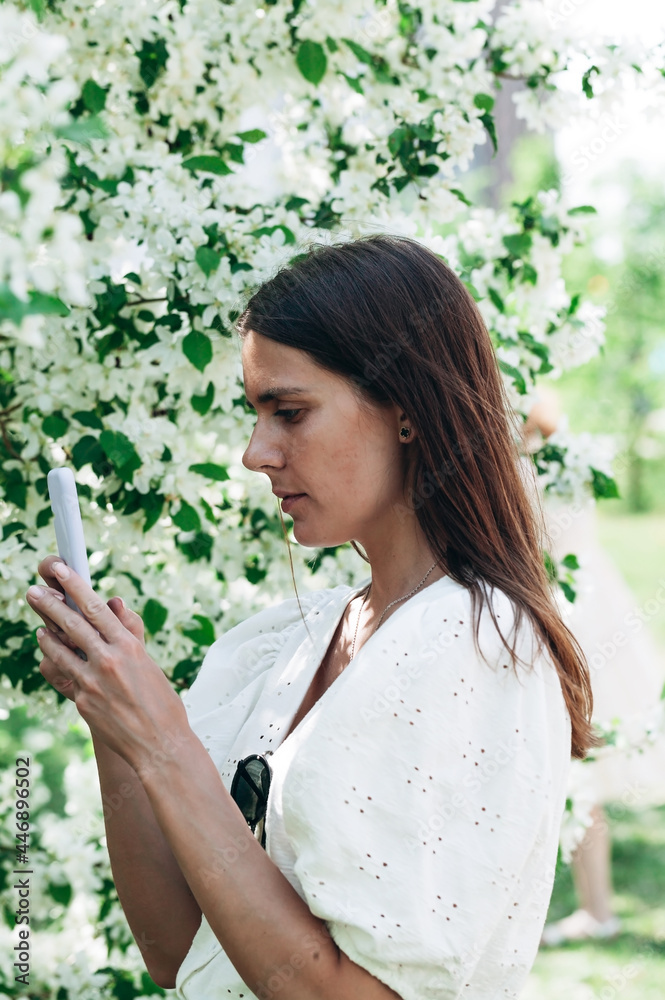 Attractive and beautiful woman on the apple tree background uses mobile technologies. Young woman caucasian ethnicity. Cheerful pretty model with phone. Female posing in white clothes. Vertical shot. Attractive and beautiful woman on the apple tree background uses mobile technologies. Young woman caucasian ethnicity. Cheerful pretty model with phone. Female posing in white clothes. Vertical shot.
