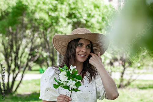 Pretty and attractive female with apple flower. Young lady in on a spring park background. Cute smiling and happy young woman in a straw hat. Beautiful model on the white blouse. Life style photo