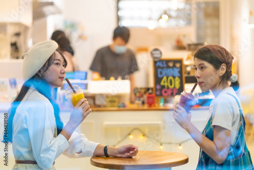 Young Asian woman friends sitting by the window in cafe with drinking fruit juice and talking together with happiness. Attractive female friendship enjoy leisure activity and meeting in the city