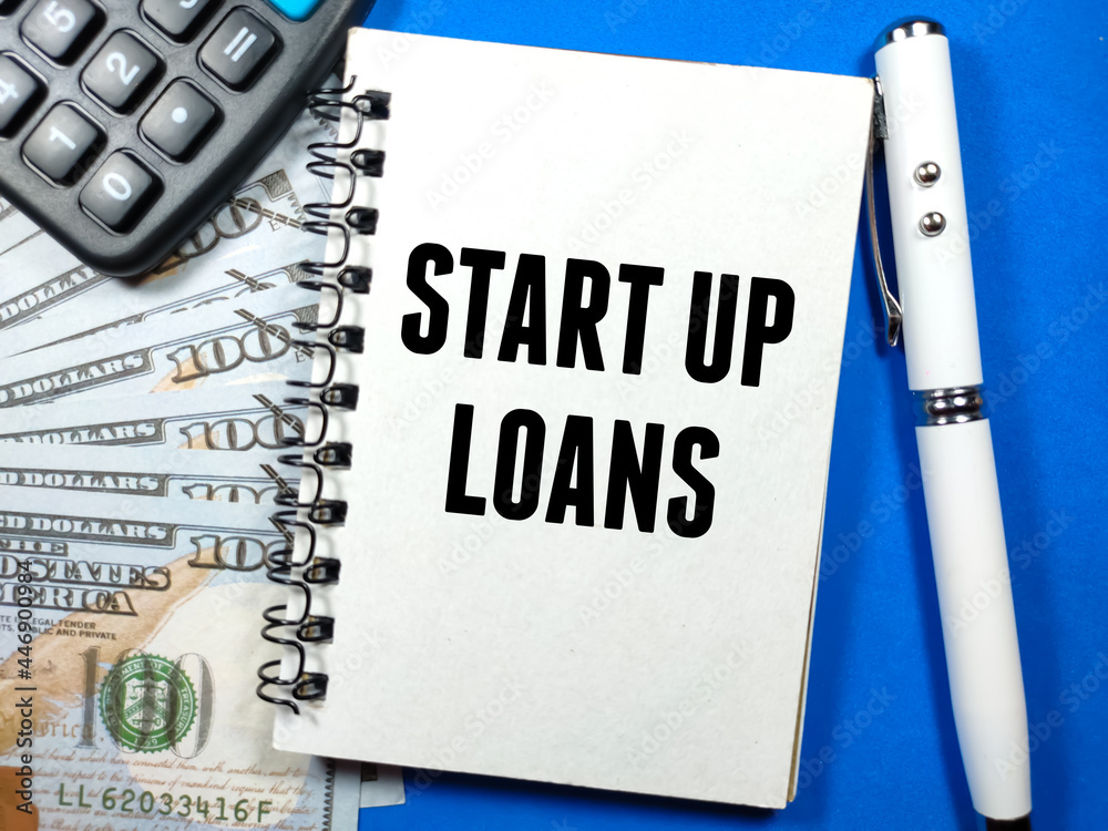 Finance concept.Text START UP LOANS writing on notebook with pen,calculator and banknote on a blue background. Finance concept.Text START UP LOANS writing on notebook with pen,calculator and banknote on a blue background.