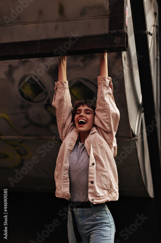 Excited brunette girl hugs on iron pipe outdoors. Stylish woman in grey t-shirt, jeans and light jacket has fun outside..