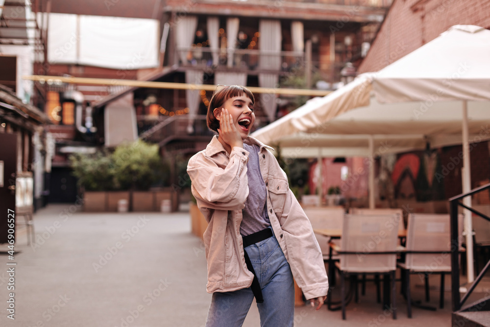 Stylish teen with brunette hairstyle in denim clothes has fun outside. Excited woman with short hair in beige jacket and jeans posing in city..