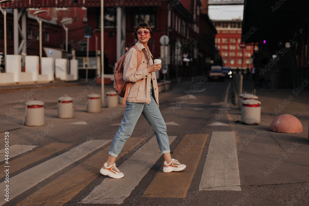 Optimistic lady in light outfit walks outside. Modern girl in jeans, sneakers, glasses and beige jacket with backpack holds cup of tea at street..