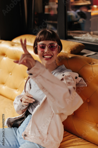 Stylish girl in pink sunglases smiles sincerely and sits on yellow couch in cafe. Pisitive lady in denim outfit smiles and shows peace sign inside..