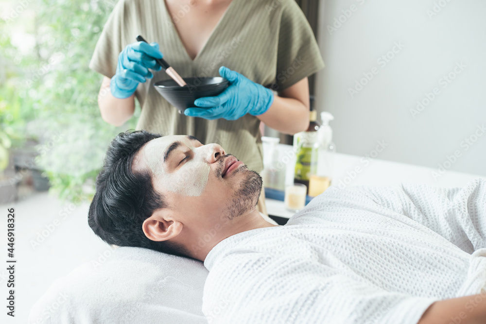 Concept facial treatment and skin rejuvenation for men. Beautician use clay mask on Asian man face. A man relaxation in spa salon. Young man lying relaxation facial face mask in spa salon.