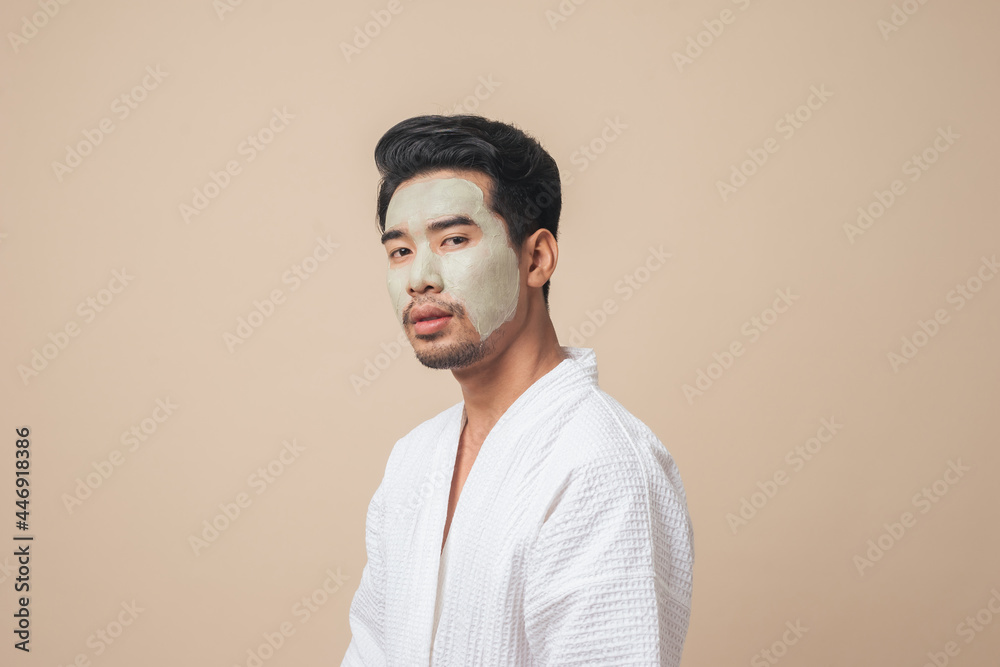 Asian man in bathrobe facial treatment and skin rejuvenation with clay mask on face in studio brown color background. - obrazy, fototapety, plakaty 