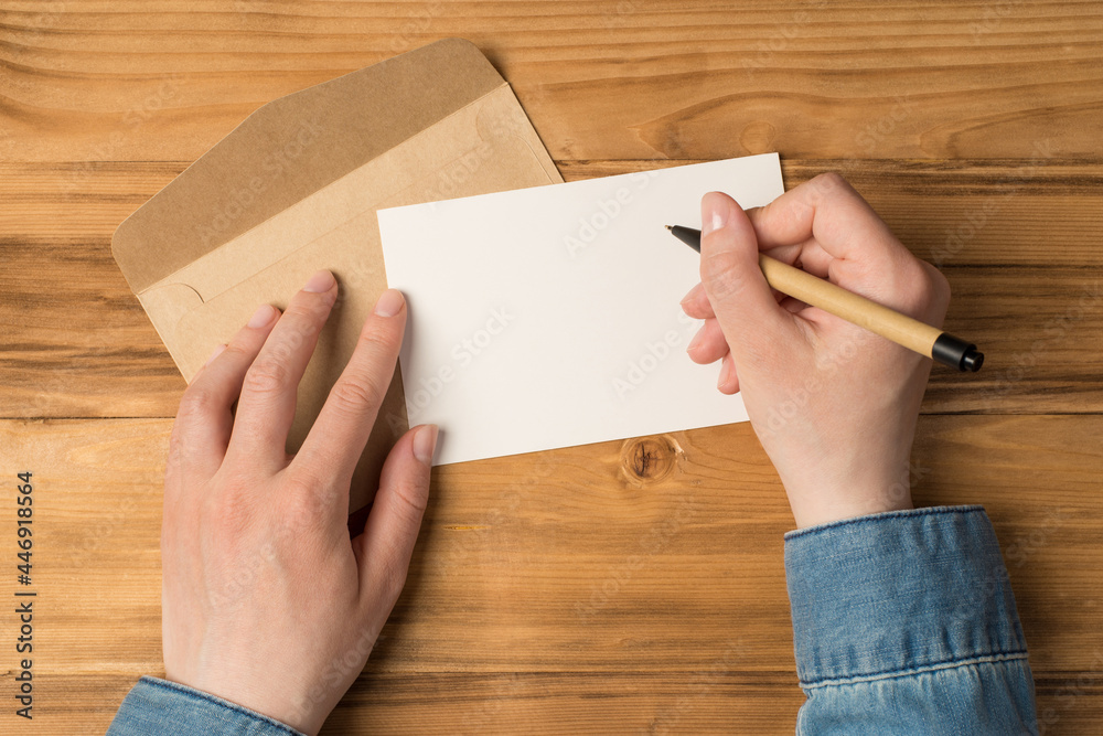 First person top view photo of hands holding pen over white card and craft paper envelope on isolated wooden table background with blank space First person top view photo of hands holding pen over white card and craft paper envelope on isolated wooden table background with blank space