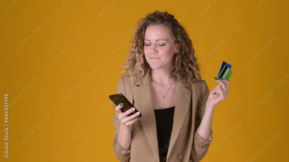 Happy young expressive girl with curly hair Using mobile cell phone hold credit bank card makes purchases online shopping isolated on yellow background. People lifestyle concept. Happy young expressive girl with curly hair Using mobile cell phone hold credit bank card makes purchases online shopping isolated on yellow background. People lifestyle concept.