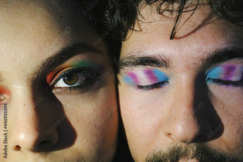 portrait of closeup faces of two lgbtq queer friends faces with rainbow pride flag and trans pride flag makeup / one eye open version - obrazy, fototapety, plakaty 