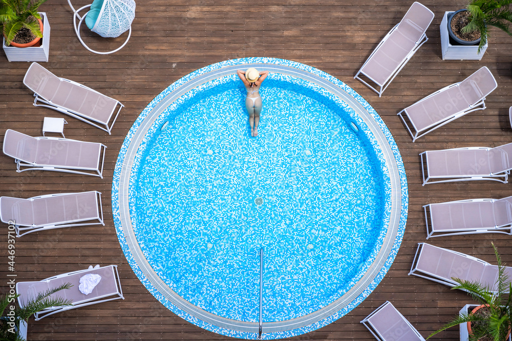 Young alone beautiful sports woman in bikini and hat is swimming in hydro massage bath on wooden terrace. Aerial drone view. Tropical summer luxury hotel