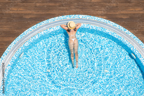 Young alone beautiful sports woman in bikini and hat is swimming in hydro massage bath on wooden terrace. Aerial drone view from above. Tropical summer luxury hotel photo